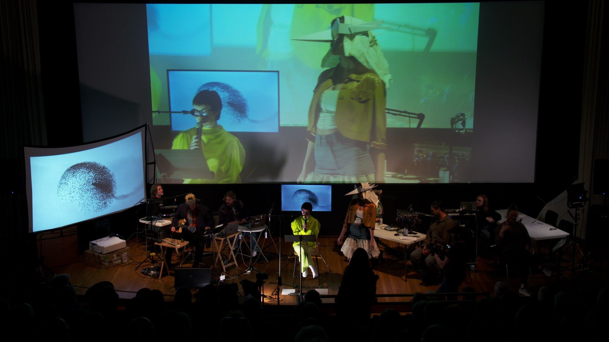 A wide view of the full stage: performers, musicians, microphones, and multiple screens. A masked performer and reader appear both onstage and in the large live projection above them.