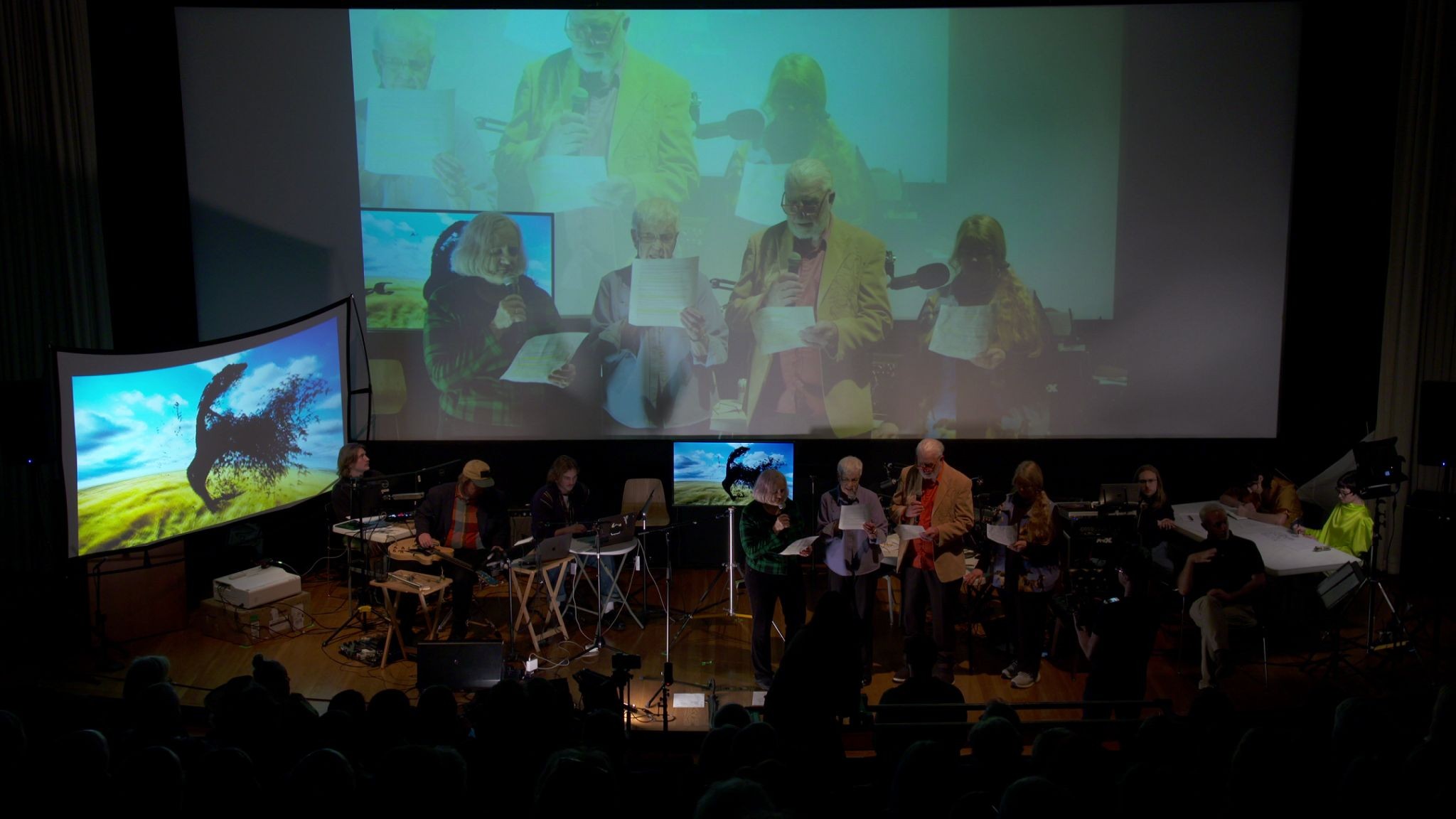 Four readers stand together with printed scripts and microphones while their live image fills the screen above them.