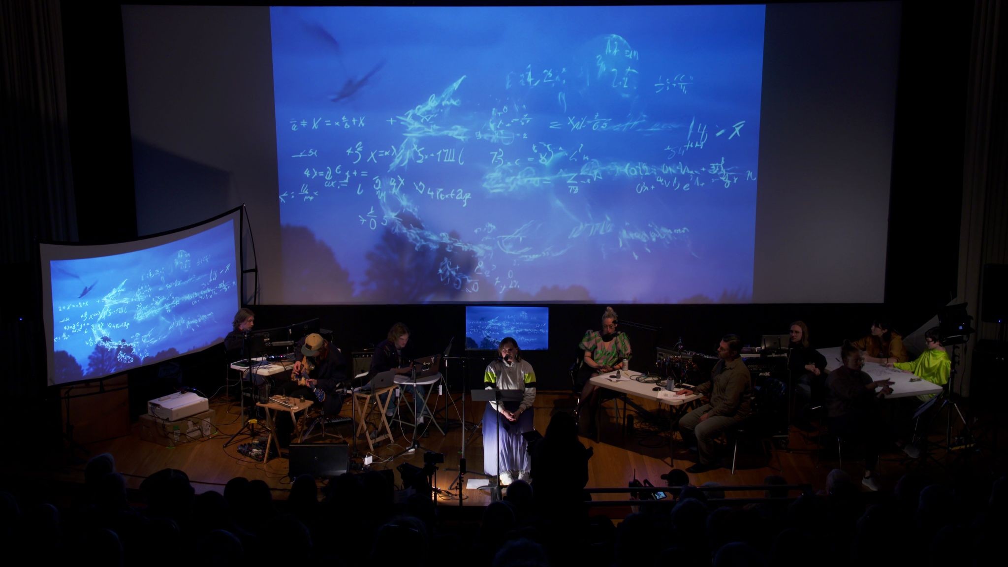 A seated reader speaks at a microphone beneath a blue projection of equations and birds. Musicians and other performers sit on either side.