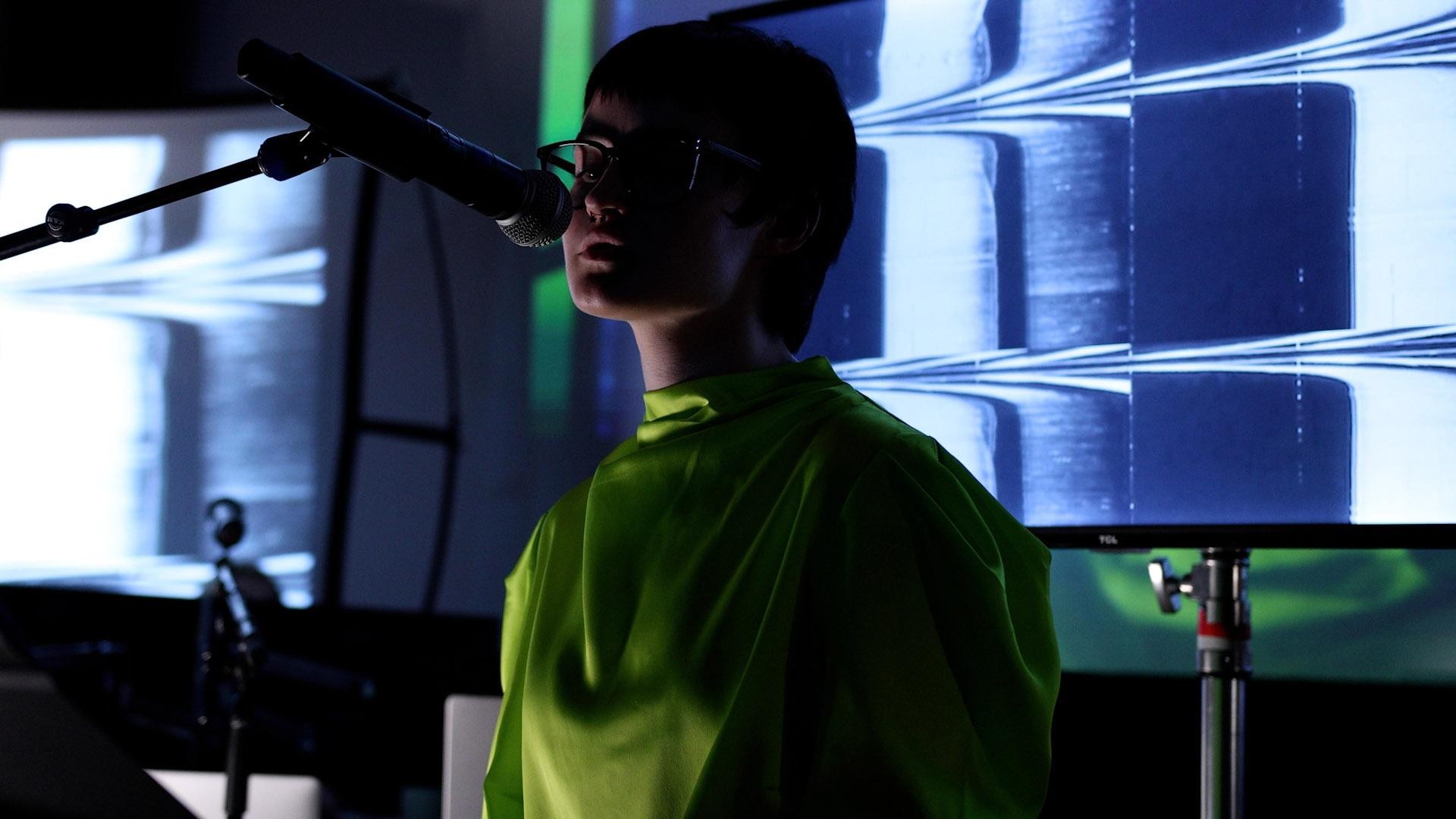 A performer in a bright green costume speaks at a microphone in front of projected geometric lines.